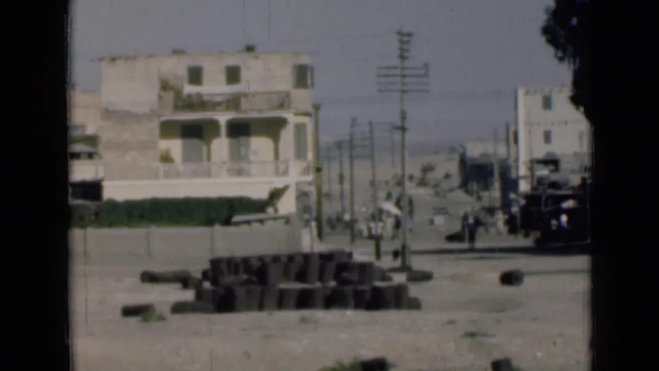 1946 Egypt home-movie frame near the Suez Canal with a pale building, roadside blocks, utility poles, pedestrians, trees, and vehicles.