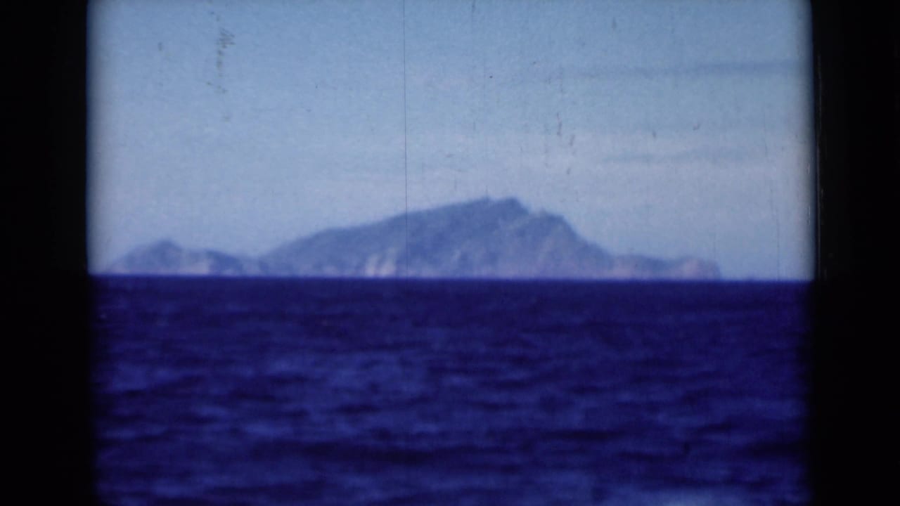 Distant mountainous landform across dark blue sea beneath a pale sky.