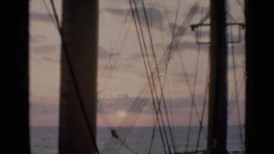 Shipboard 8mm sunset frame with pale pink sky, distant horizon, silhouetted rigging, and dark vertical vessel structures.