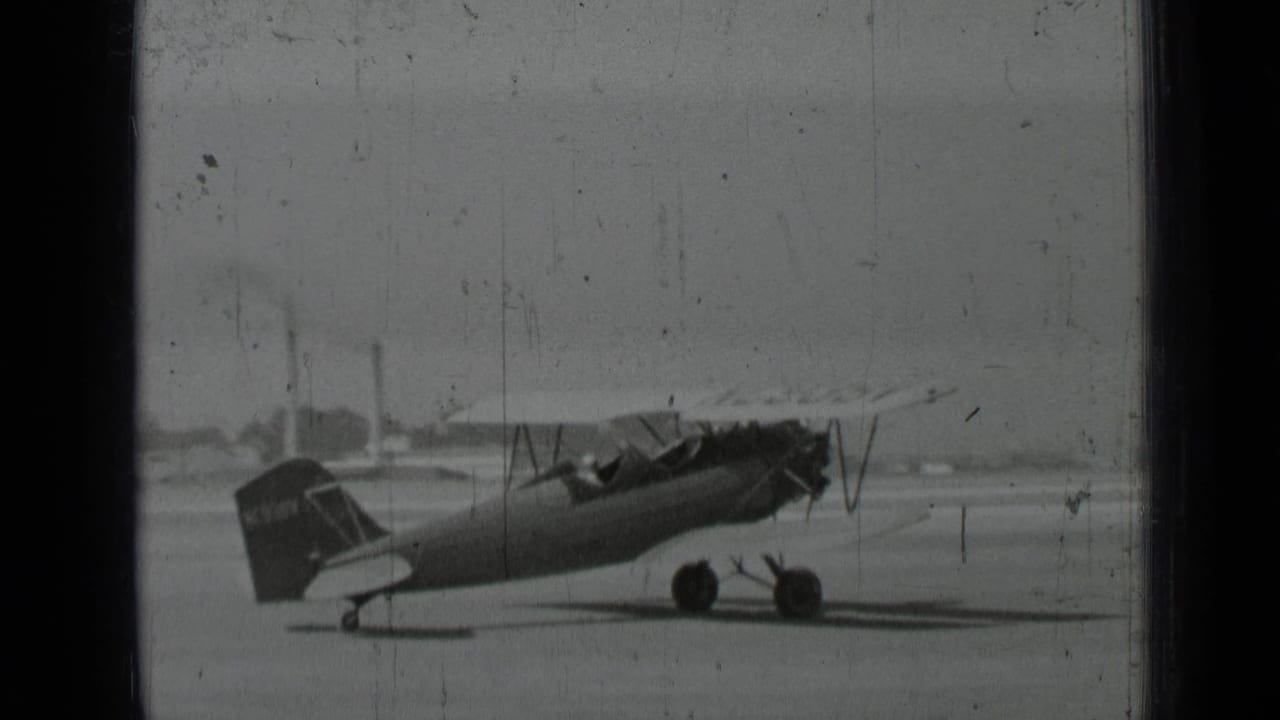 Biplane parked on an open tarmac in black-and-white home-movie footage.