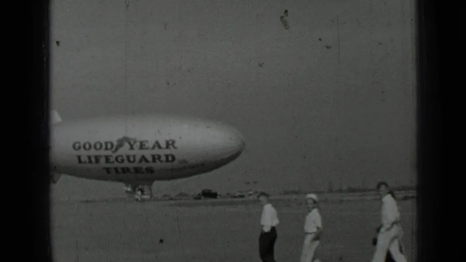 1937 San Francisco-area frame with a Goodyear Lifeguard Tires blimp behind several people walking on the tarmac.