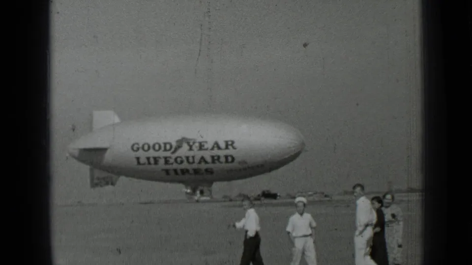 Home-movie frame from the 1937 San Francisco clip showing spectators near a Goodyear Lifeguard Tires blimp on an airfield.