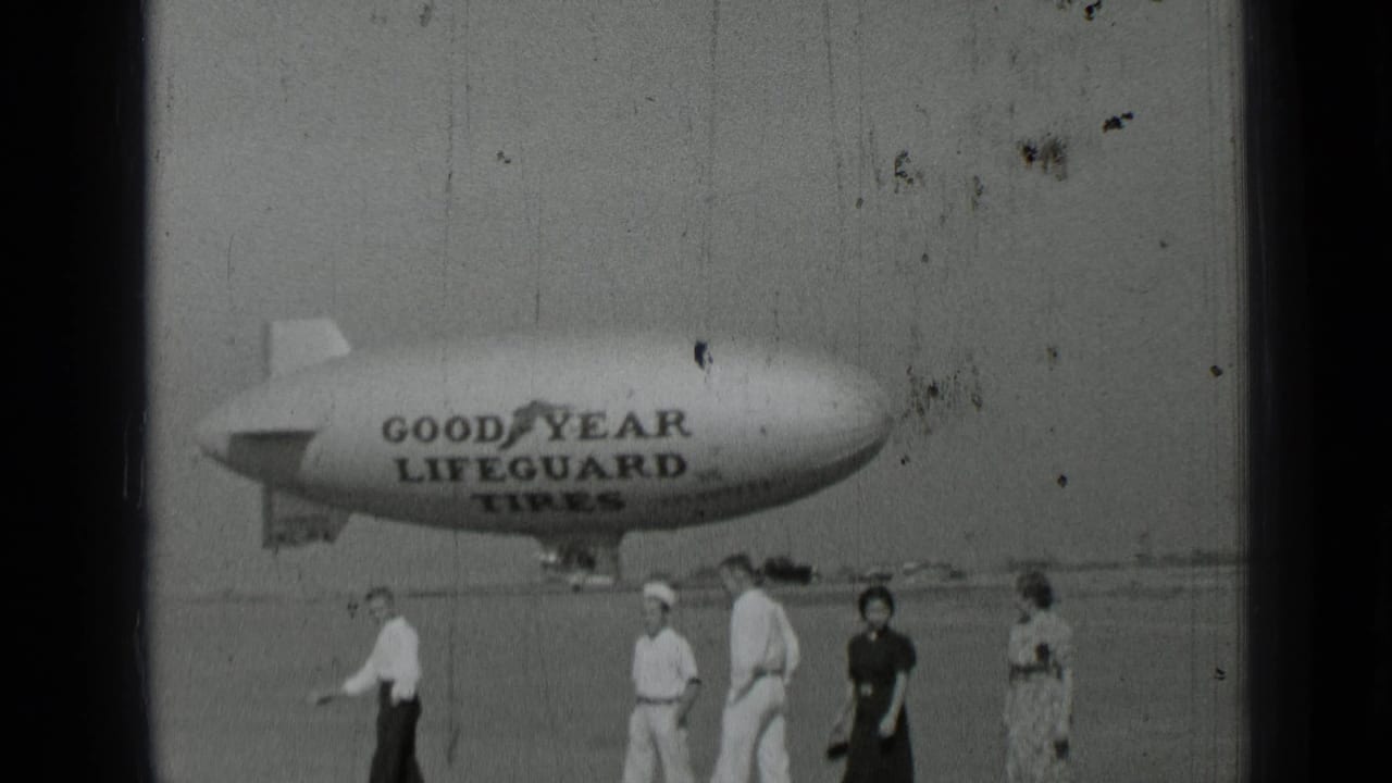 Adults walk across an open field with a Goodyear Lifeguard Tires blimp behind them.