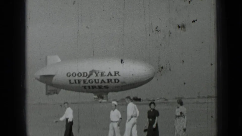 1937 San Francisco home-movie frame showing adults walking in front of a Goodyear Lifeguard Tires blimp on an open field.