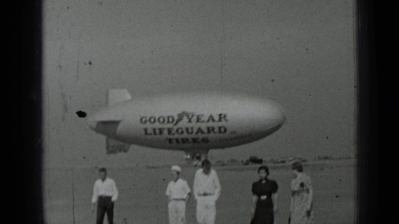 Several adults stand and walk in front of a Goodyear Lifeguard Tires blimp.
