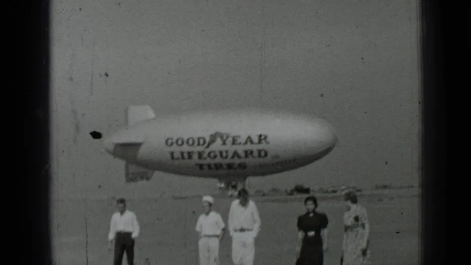 1937 San Francisco 8mm frame with several adults near a large Goodyear Lifeguard Tires blimp on a flat field.
