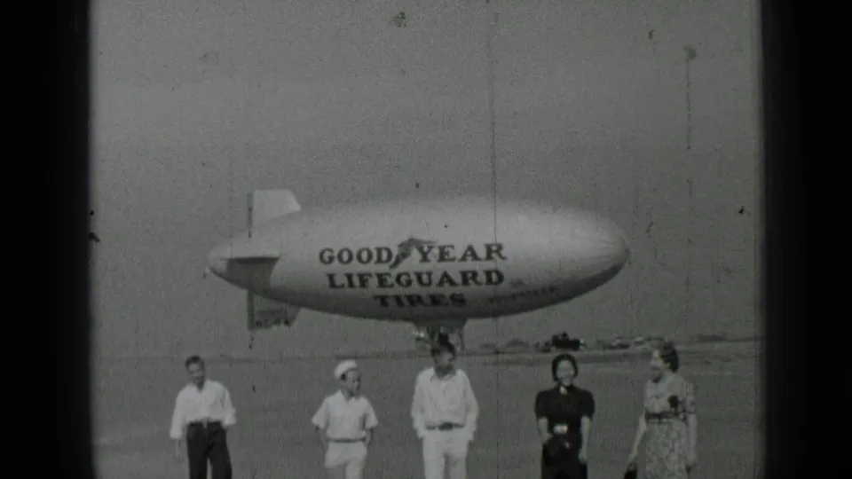 1937 San Francisco home-movie frame showing adults arranged below a Goodyear Lifeguard Tires blimp in a wide open field.