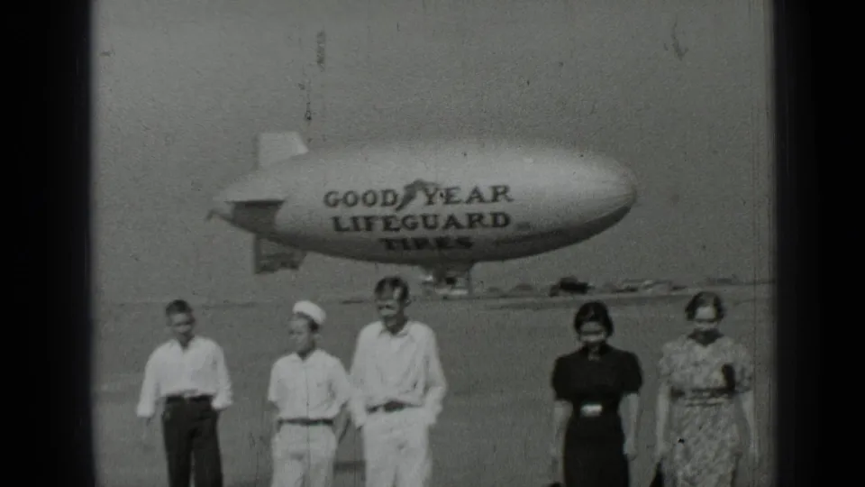 Five people in period clothing walk toward the camera on a San Francisco airfield with the Goodyear blimp behind them.