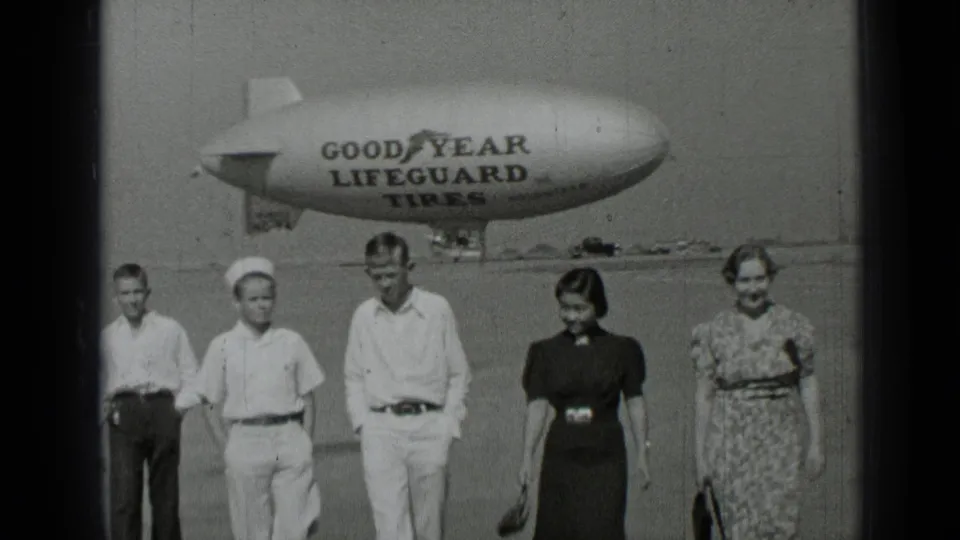 The group walks beneath the visible Goodyear Lifeguard Tires blimp lettering in black-and-white 1937 airfield footage.