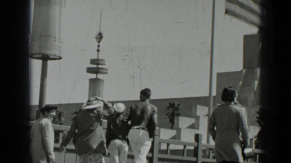 1937 San Francisco pedestrians seen from behind on a public walkway with railings, lampposts, and distant palms.