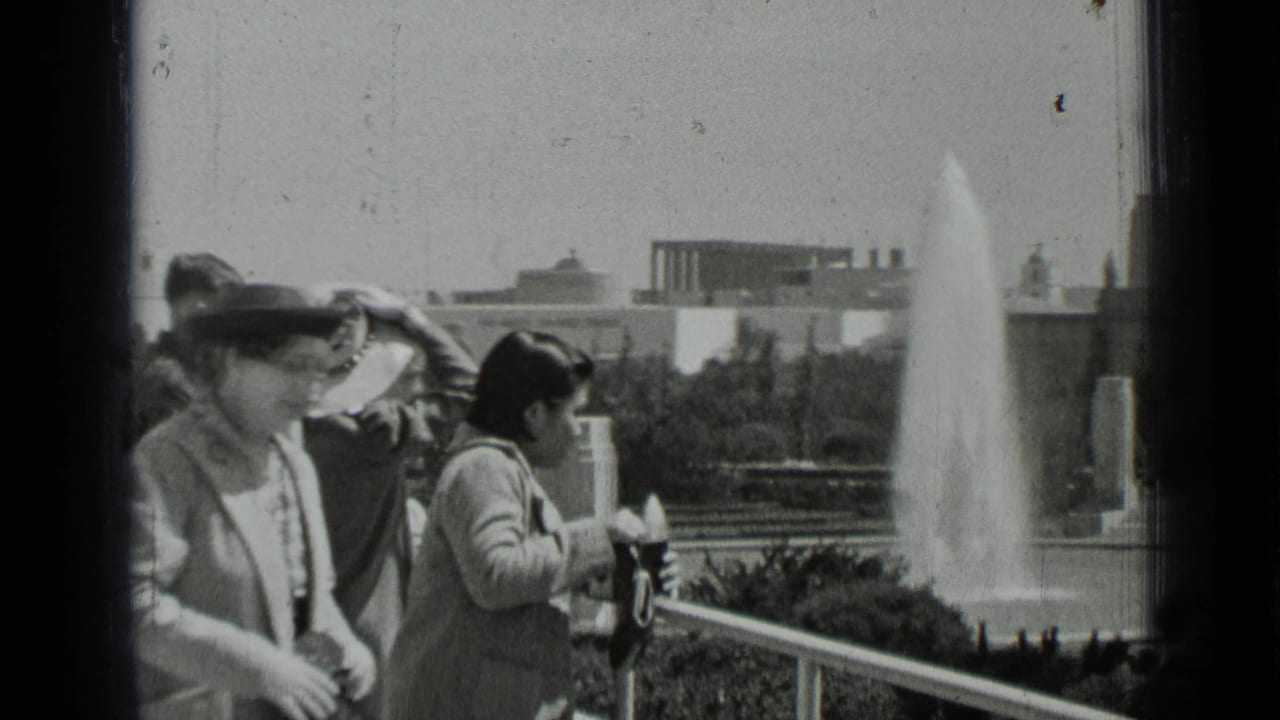 Visitors stand by a balcony rail overlooking a large fountain and distant buildings.