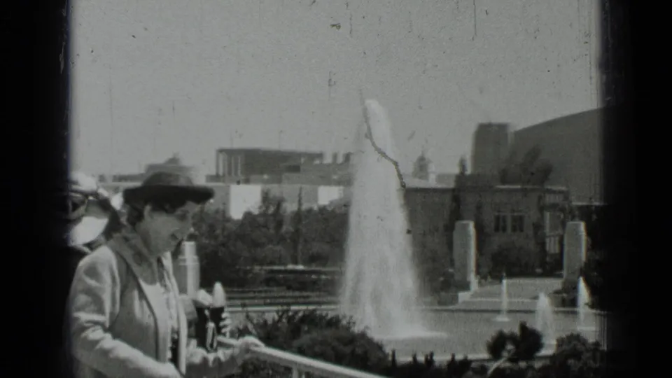 1937 San Francisco frame of a woman in a dark hat and light coat near a balcony rail, with a fountain and buildings in the background.
