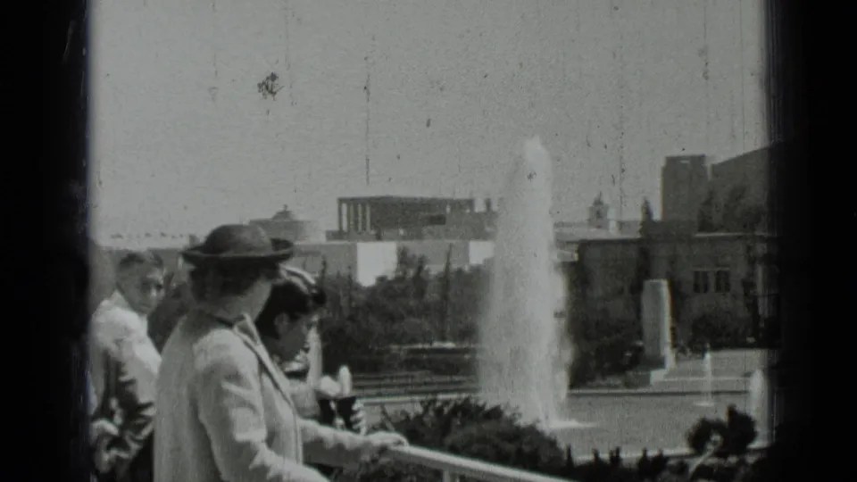 1937 San Francisco home-movie still with visitors at a balcony rail, a woman in a dark hat, and a tall fountain plume in the background.