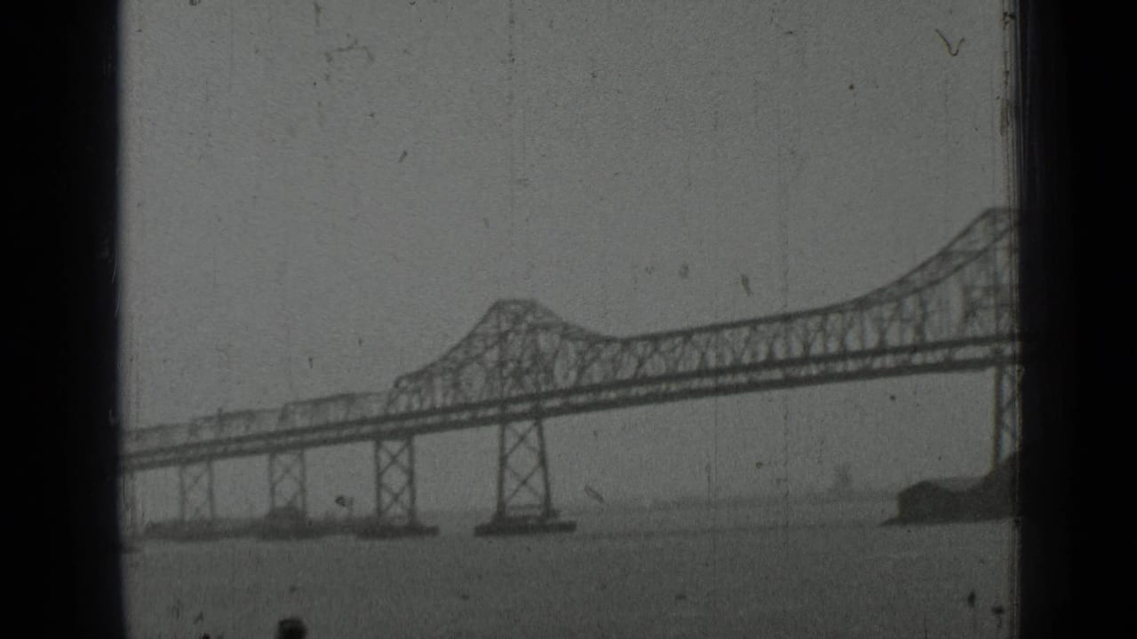 Distant steel truss bridge spanning water, framed by dark film borders.