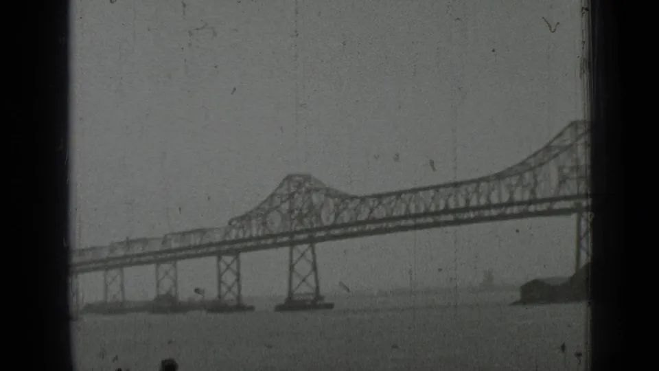 1937 San Francisco home-movie frame showing a distant steel truss bridge across bay water with soft focus and visible film wear.