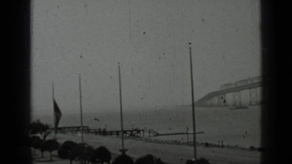 1937 San Francisco waterfront promenade with flagpoles, small trees, railings, and the bridge receding across the water.
