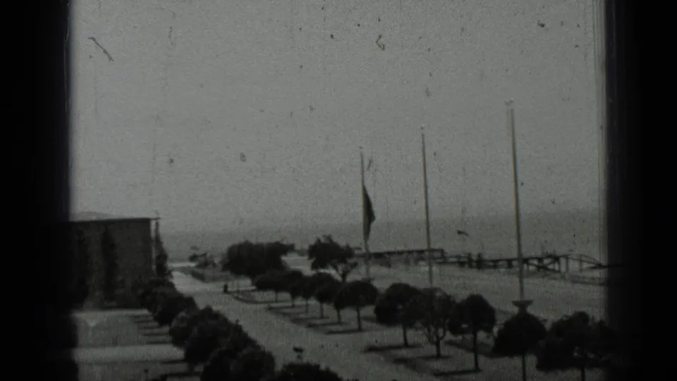 San Francisco waterfront promenade in 1937, showing trimmed trees, flagpoles, railings, water, and a building edge in soft 8mm focus.