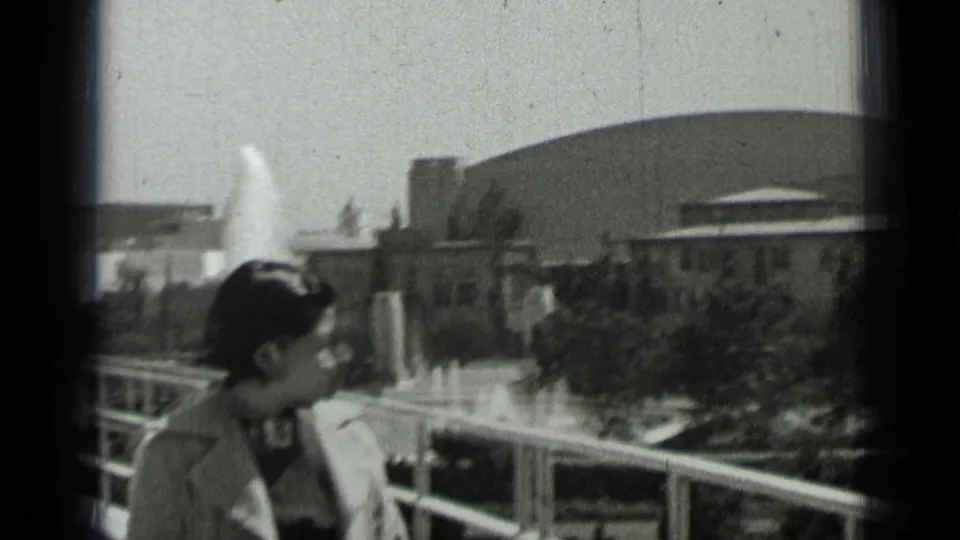 A woman in period attire stands near a balcony railing as a fountain sprays before a domed San Francisco building.