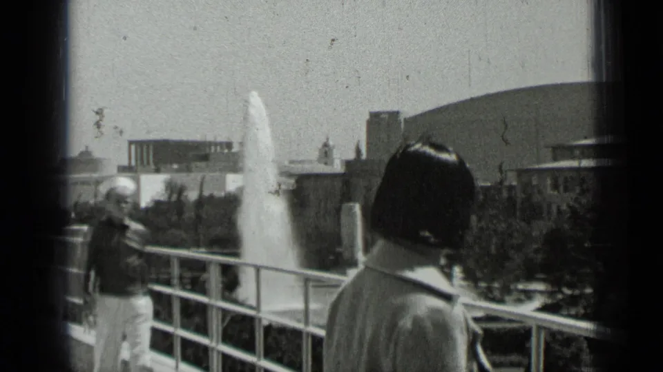 Two adult visitors stand or pass near a balcony railing, with fountain spray and a domed San Francisco building in the background.