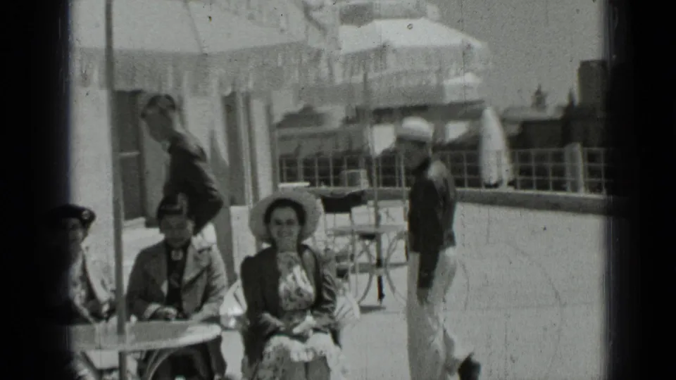Adults in period dress sit at an outdoor table in San Francisco while a sailor in uniform stands near the railing.