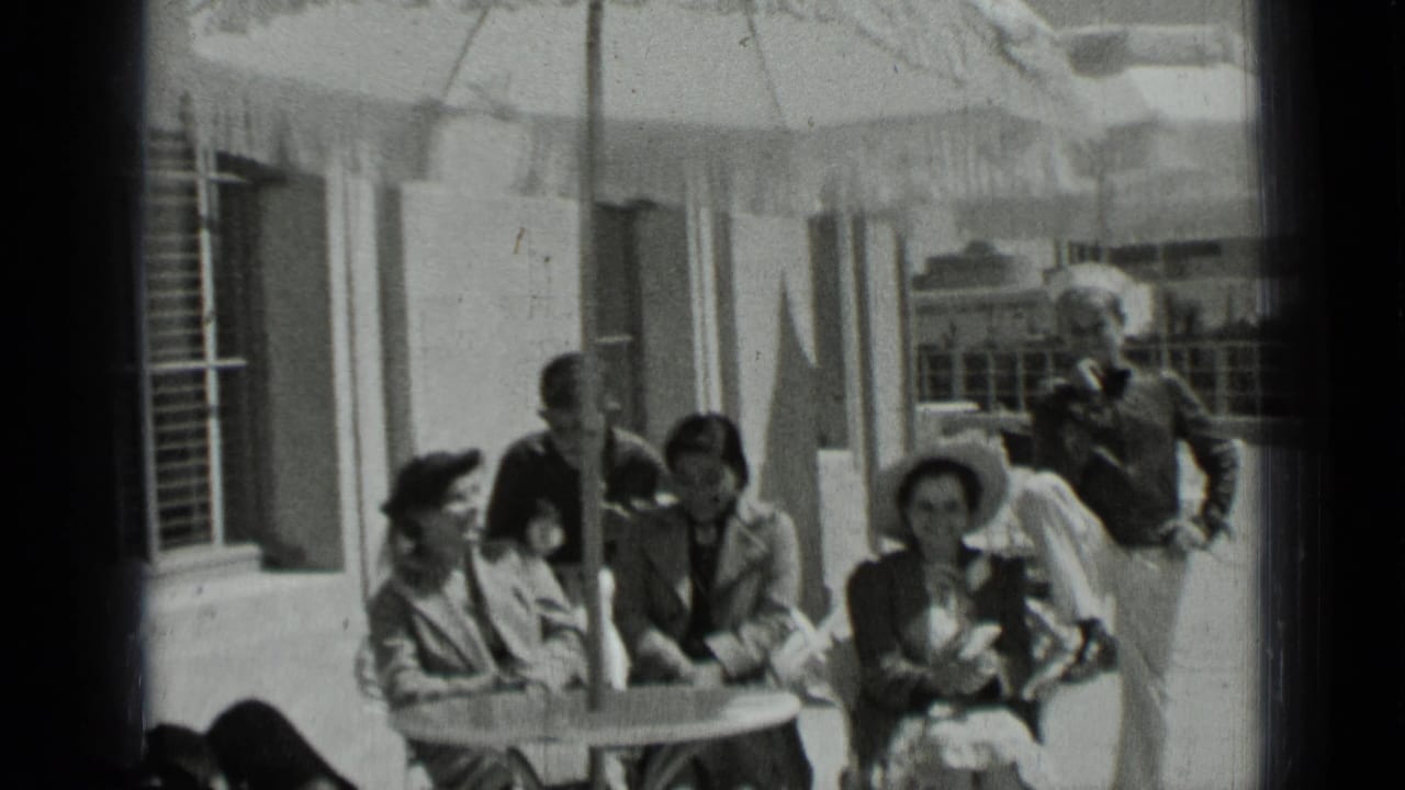 1937 San Francisco home-movie frame of women in hats seated at a patio table while men stand nearby under a fringed umbrella.