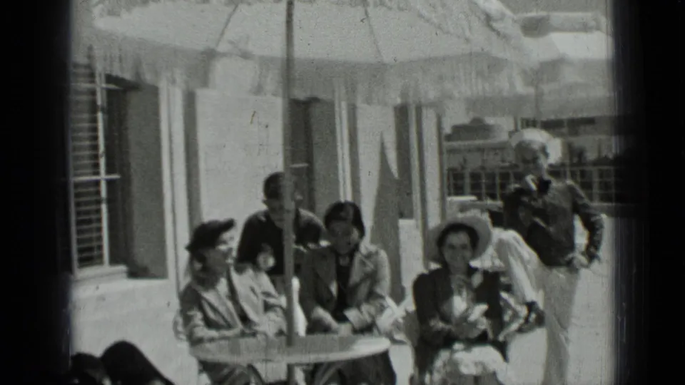 1937 San Francisco home-movie frame of women in hats seated at a patio table while men stand nearby under a fringed umbrella.