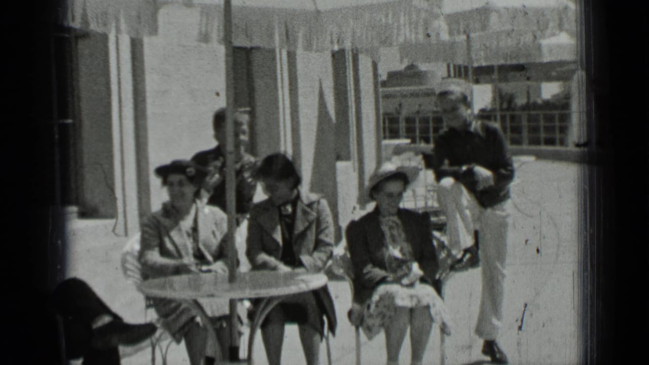 Three women sit at a patio table while a man in light trousers leans near the railing.