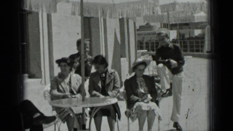 Women in period coats and hats sit at a small outdoor table in San Francisco, with a man standing beside them on a sunny deck.