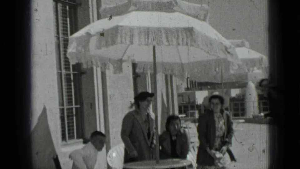 A 1937 San Francisco outdoor gathering continues as adults sit and stand under a large fringed umbrella near a building wall.