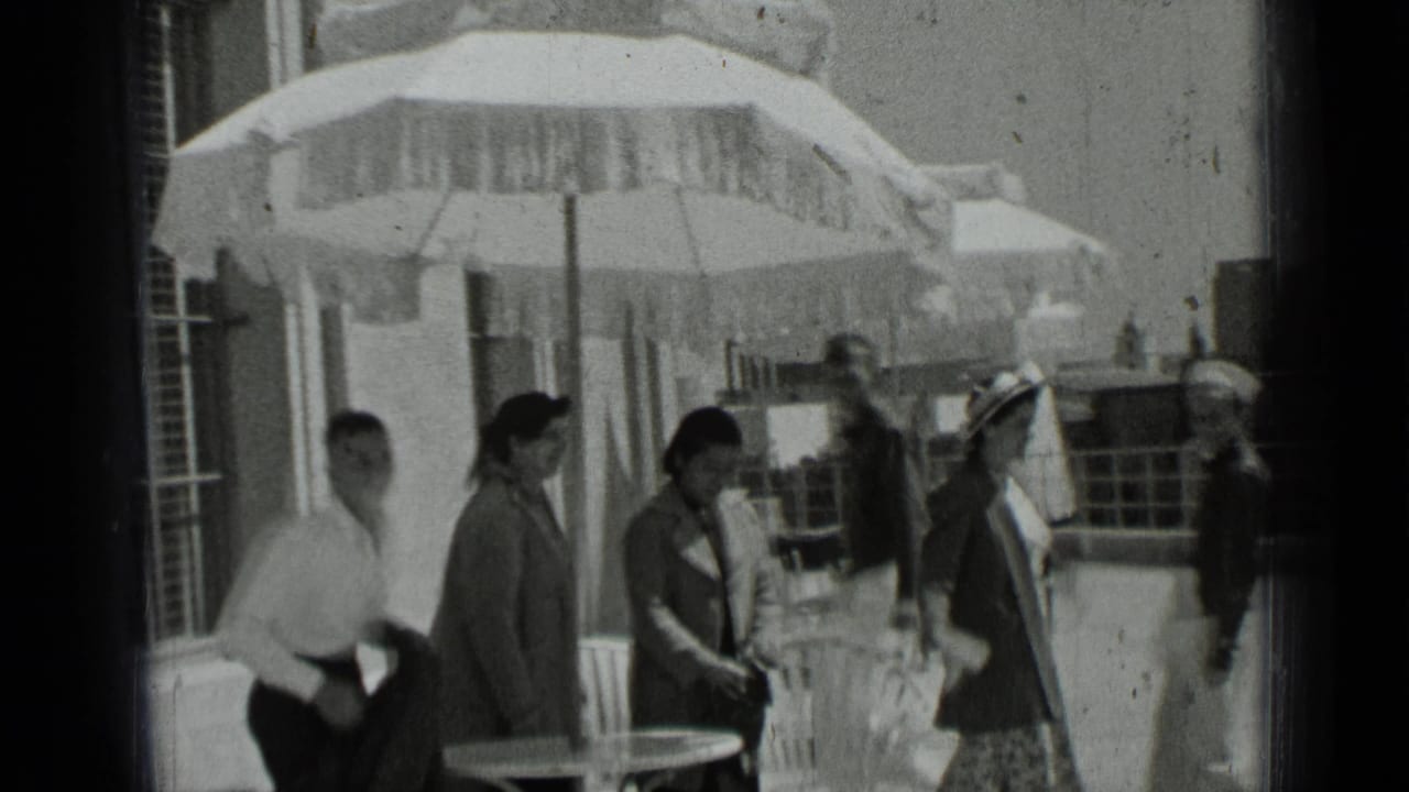 Young adults in 1930s dress walk through an outdoor patio area in San Francisco, with umbrellas, tables, and railings behind them.