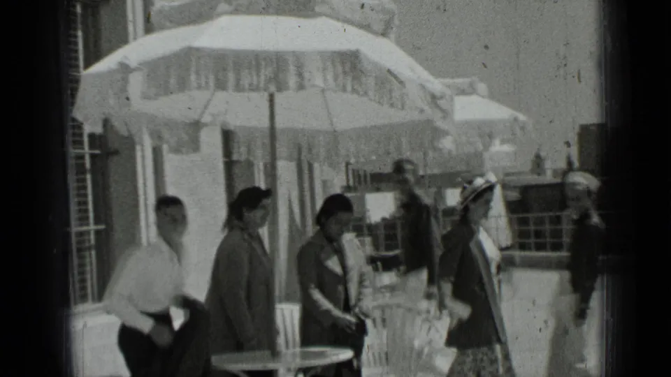 Young adults in 1930s dress walk through an outdoor patio area in San Francisco, with umbrellas, tables, and railings behind them.