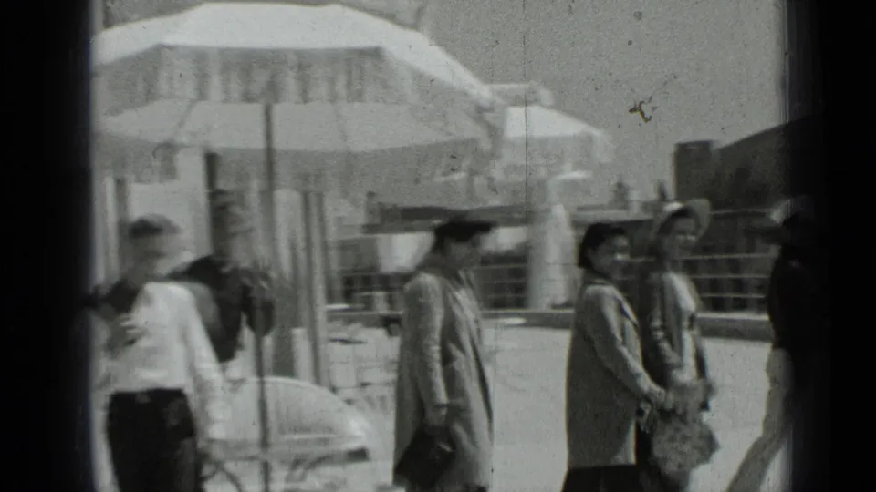 Visitors in hats and light jackets move along a sunlit San Francisco patio, with umbrellas, chairs, and a railing visible in the background.