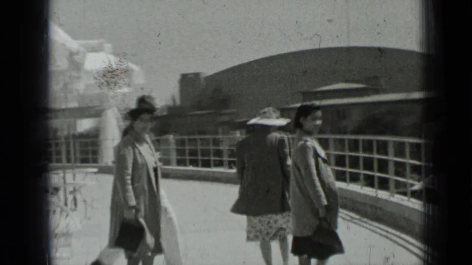 Three adults in period clothing walk along a curved railing in San Francisco, with a fountain spray and outdoor seating nearby.