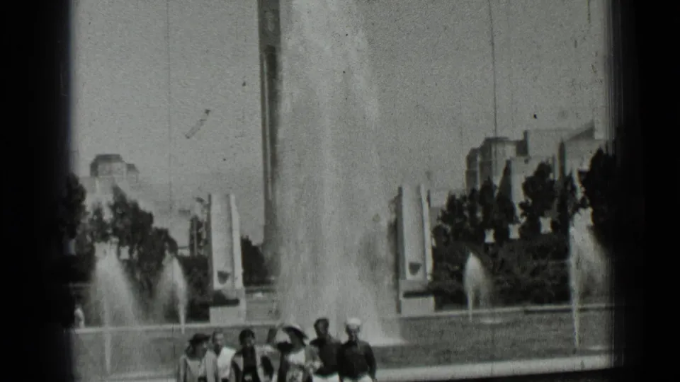 A large public fountain with tall water jets rises behind a small group of people in 1937 San Francisco home-movie footage.
