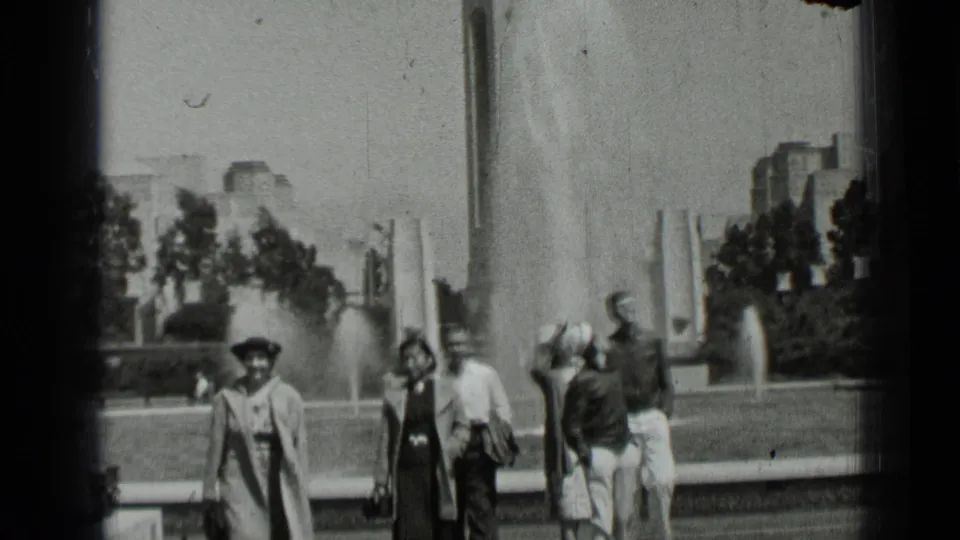 1937 San Francisco home-movie frame showing pedestrians in period dress near a fountain and trees.