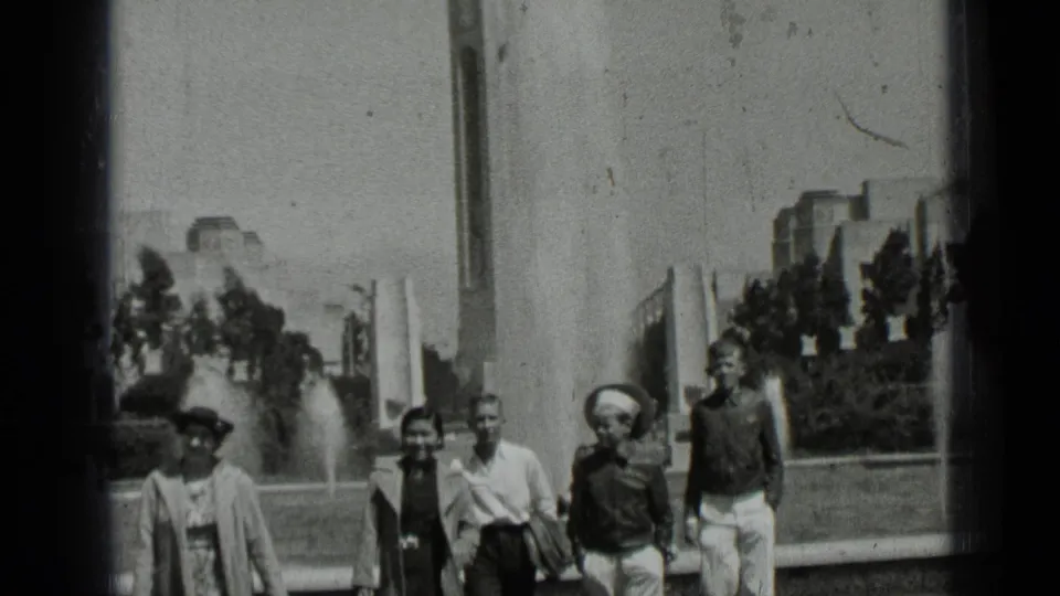 1937 San Francisco park visitors walk past a fountain, with flags, trees, and distant buildings behind them.