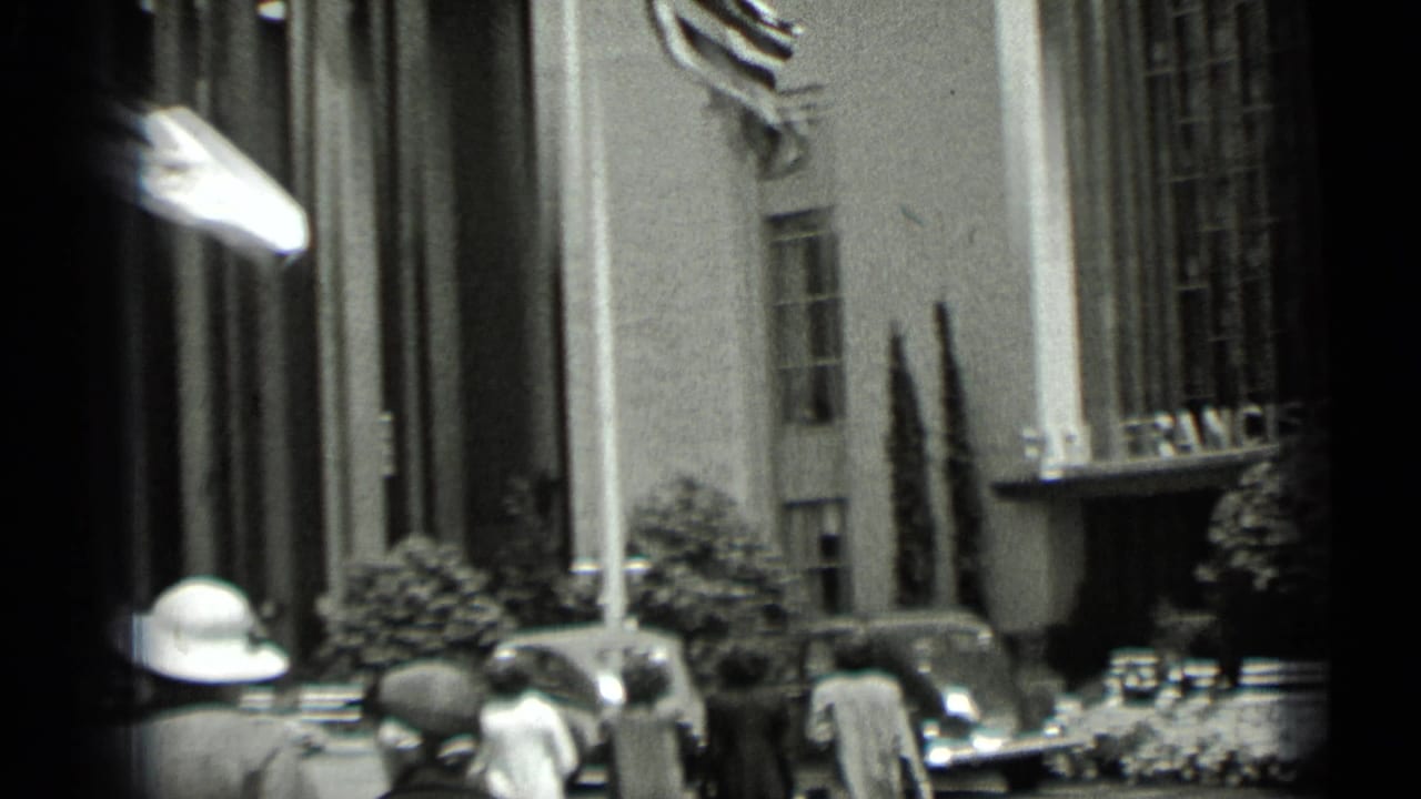 People and parked cars appear outside a tall building entrance with a flag overhead.