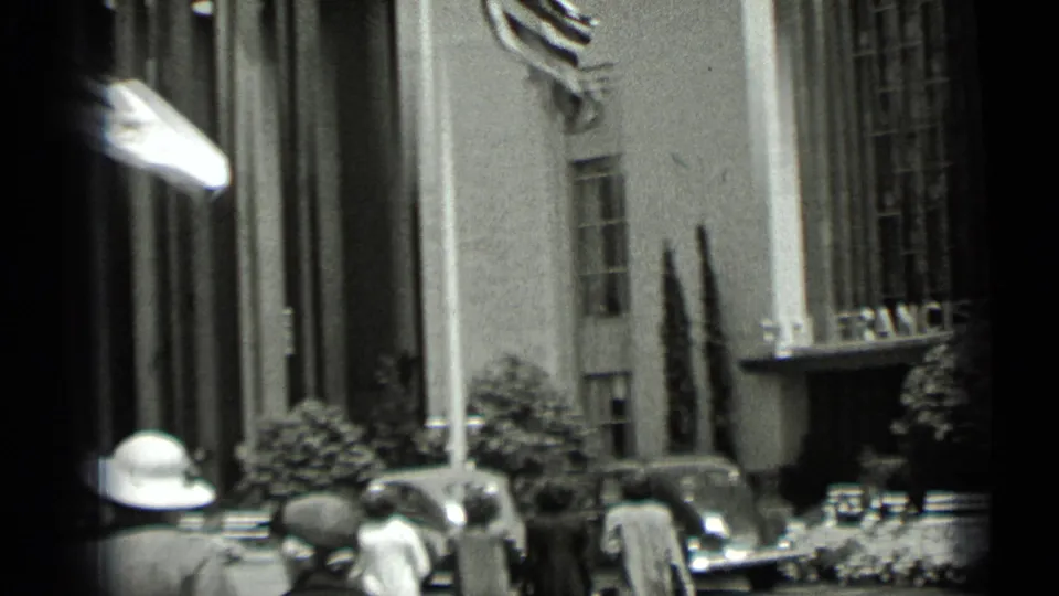 Pedestrians in period clothing and parked cars stand outside a San Francisco building entrance, with a flag moving above the street.