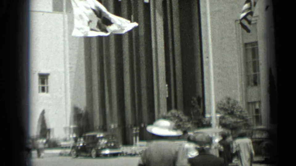 San Francisco street view from 1937 showing a flag, people in hats, parked automobiles, and a tall columned building facade.