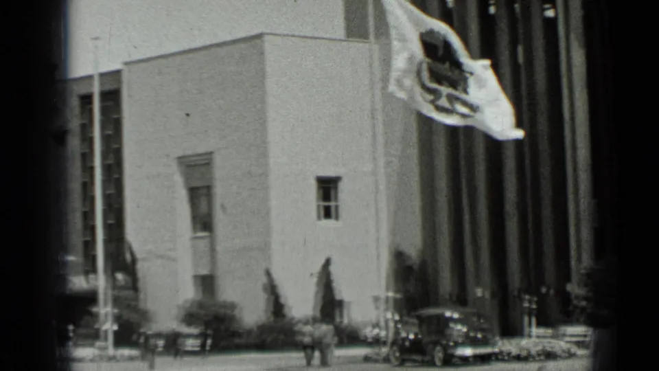 Wide 1937 San Francisco frame showing the corner of a large civic-looking building, flags, pedestrians, and parked cars near the entrance.