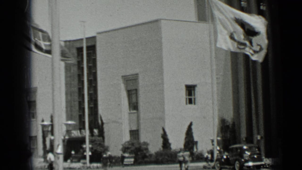 1937 San Francisco street view showing a modernist building facade with flags, small pedestrians, and a dark passenger car near the curb.