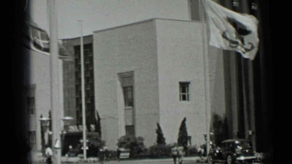1937 San Francisco street view showing a modernist building facade with flags, small pedestrians, and a dark passenger car near the curb.