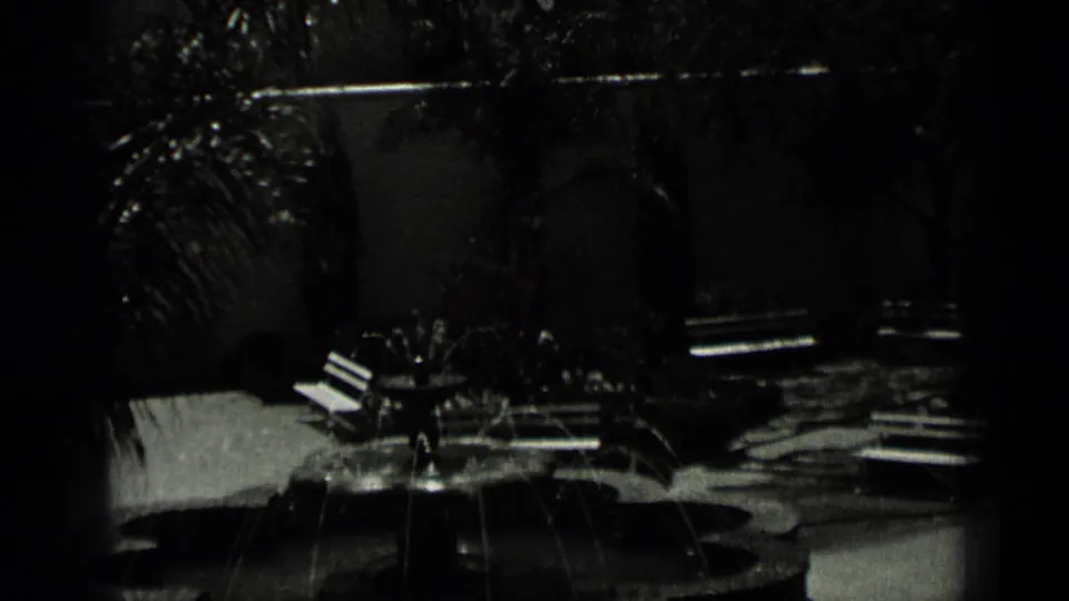 Shaded San Francisco courtyard scene from 1937 footage, with benches, planting beds, a small fountain, and a dark building wall.