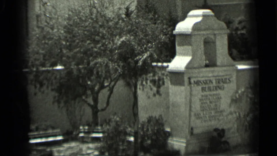 1937 San Francisco home-movie frame showing the Mission Trails Building monument near benches, trees, shrubs, and a stucco courtyard wall.