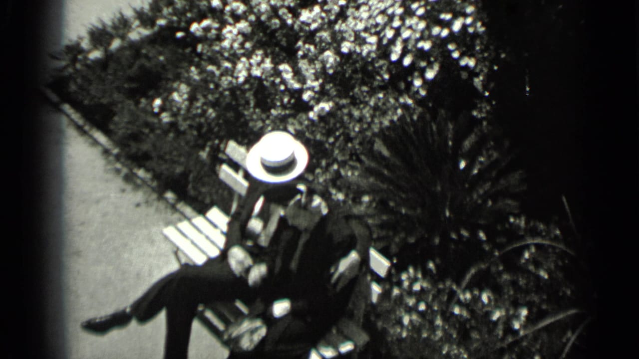 1937 San Francisco home-movie frame of a formally dressed couple sitting on a wooden bench in a planted courtyard.