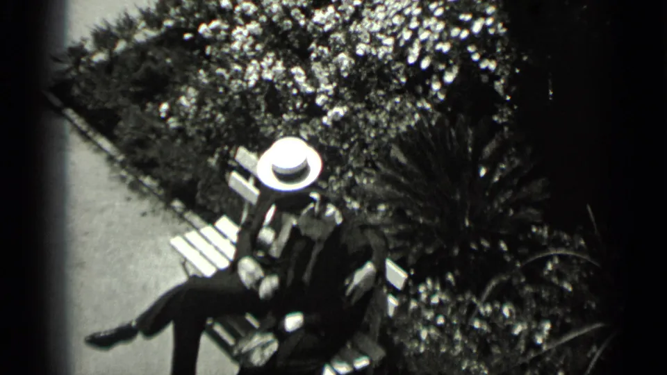 1937 San Francisco home-movie frame of a formally dressed couple sitting on a wooden bench in a planted courtyard.