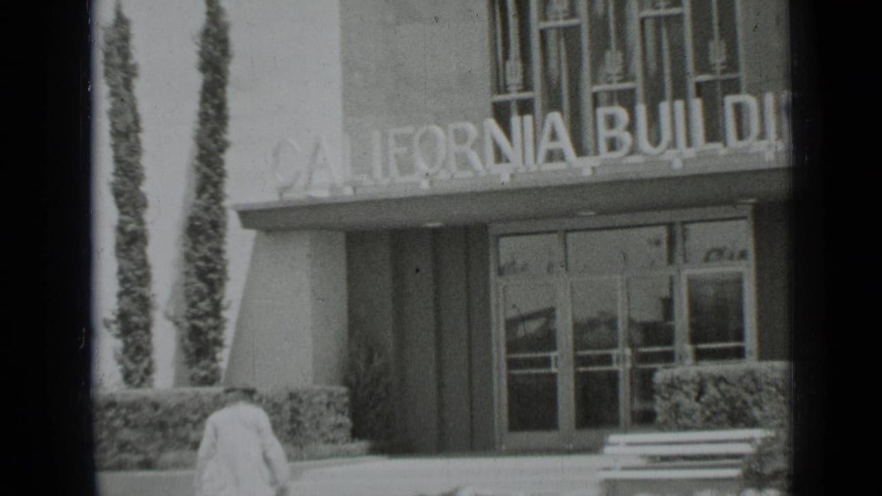 1937 San Francisco 8mm frame showing a woman approaching the California Building entrance, with glass doors, hedges, and a bench visible.