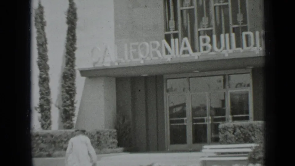 1937 San Francisco 8mm frame showing a woman approaching the California Building entrance, with glass doors, hedges, and a bench visible.