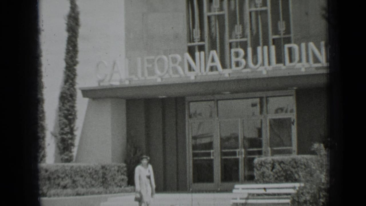 California Building entrance with sign, glass doors, hedges, bench and a pedestrian.