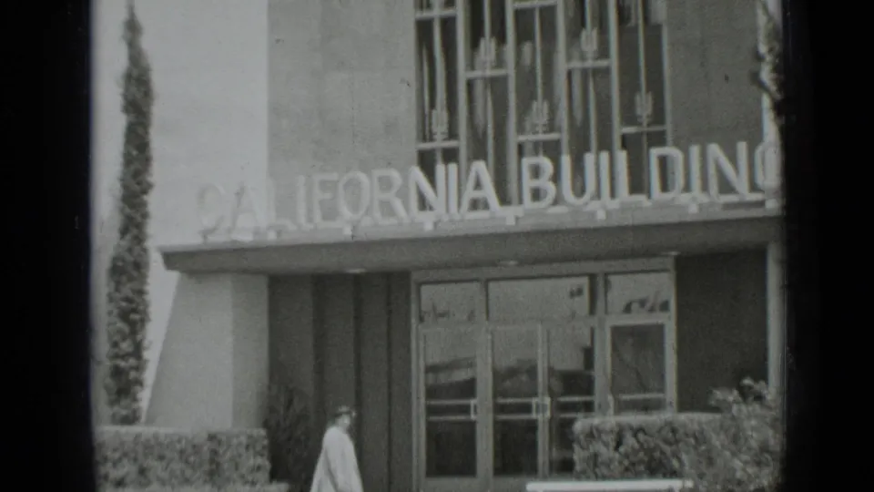 Black-and-white 1937 San Francisco frame of the California Building entrance with bold lettering and a passerby near the hedges.